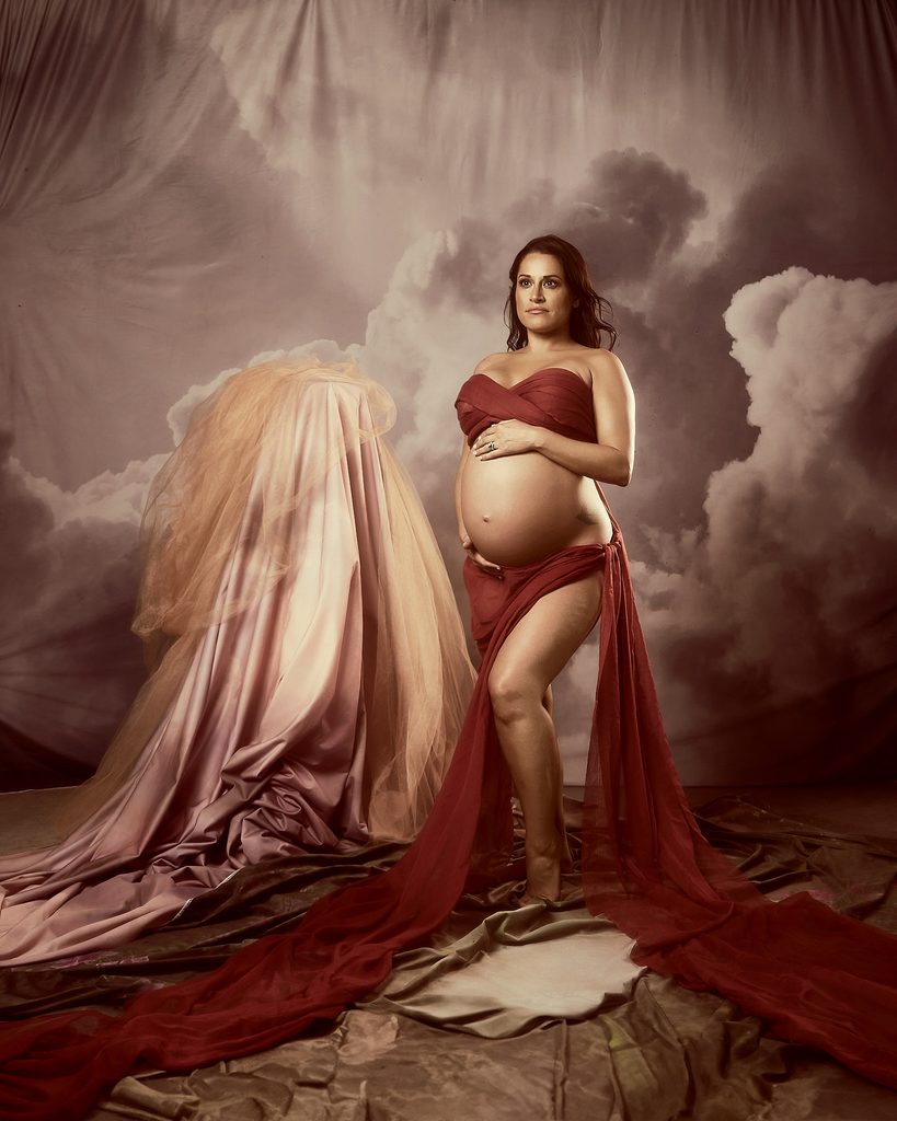 expecting pregnancy woman is holding her baby bump belly while surrounded by fabrics in a reminiscent of a renaissance painting